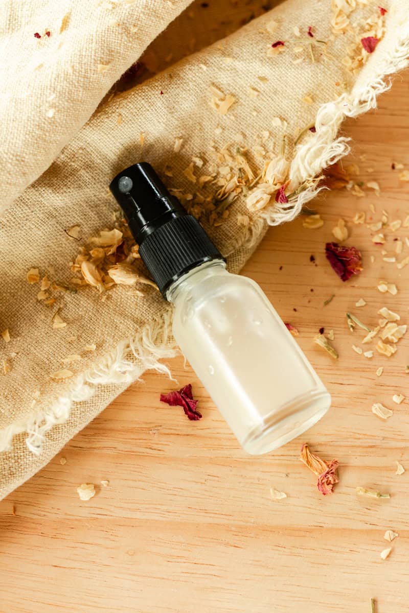 Clear glass spray bottle filled with a homemade ylang ylang spray perfume. Bottle lies on a table with burlap and dried flower petals.