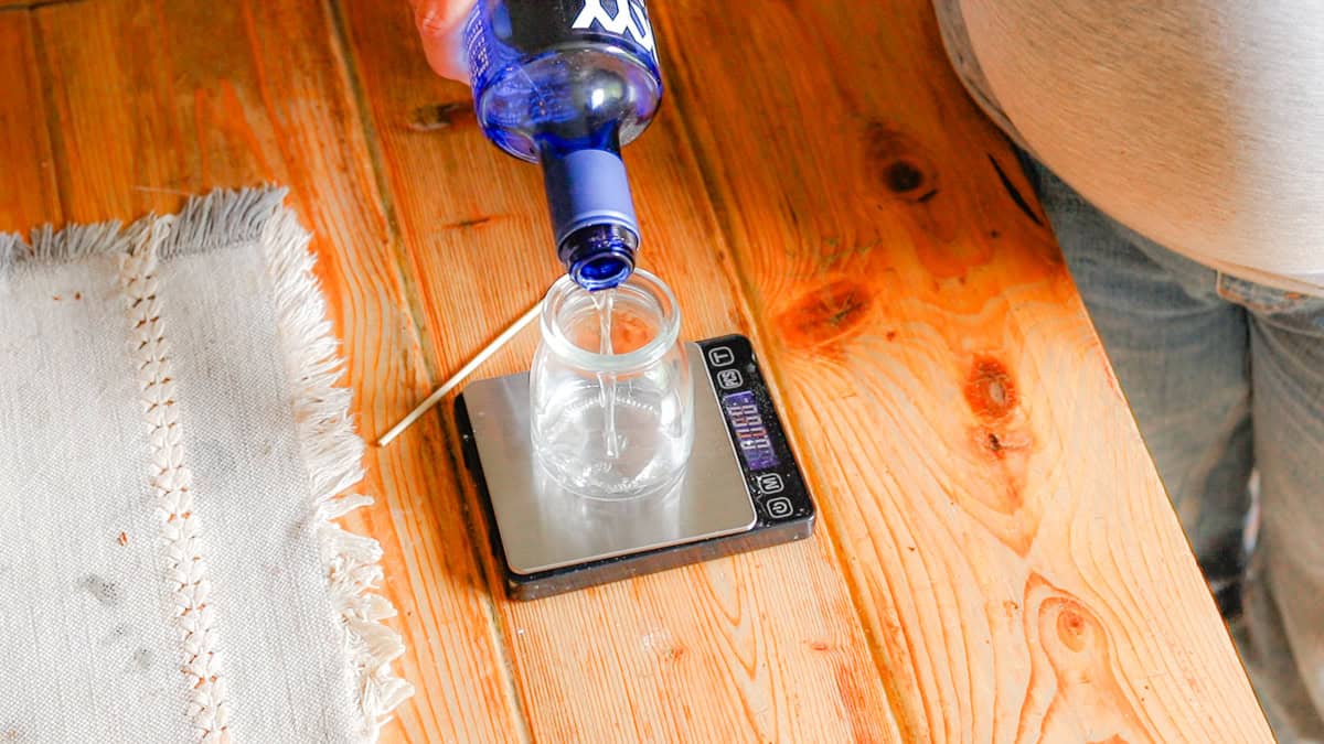 Vodka is poured into a glass jar sitting on a scale. The scale and jar sit on a wooden table with a linen table runner.