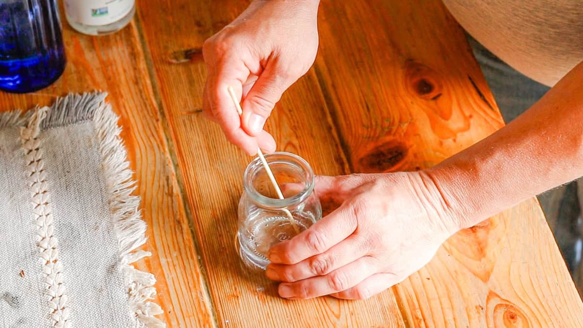 A hand is mixing ingredients in a glass jar for a homemade perfume recipe. The glass jar sits on a wooden table with a linen table runner.