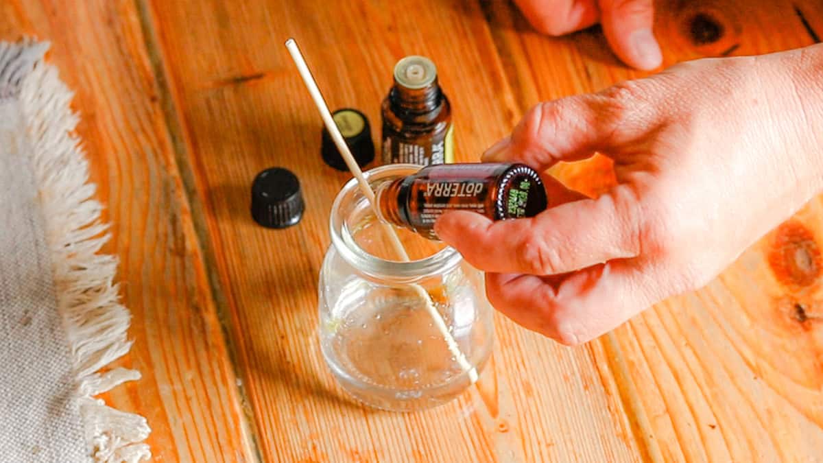 A hand is dropping essential oils into a glass jar for a homemade perfume recipe. The glass jar sits on a wooden table with a linen table runner.