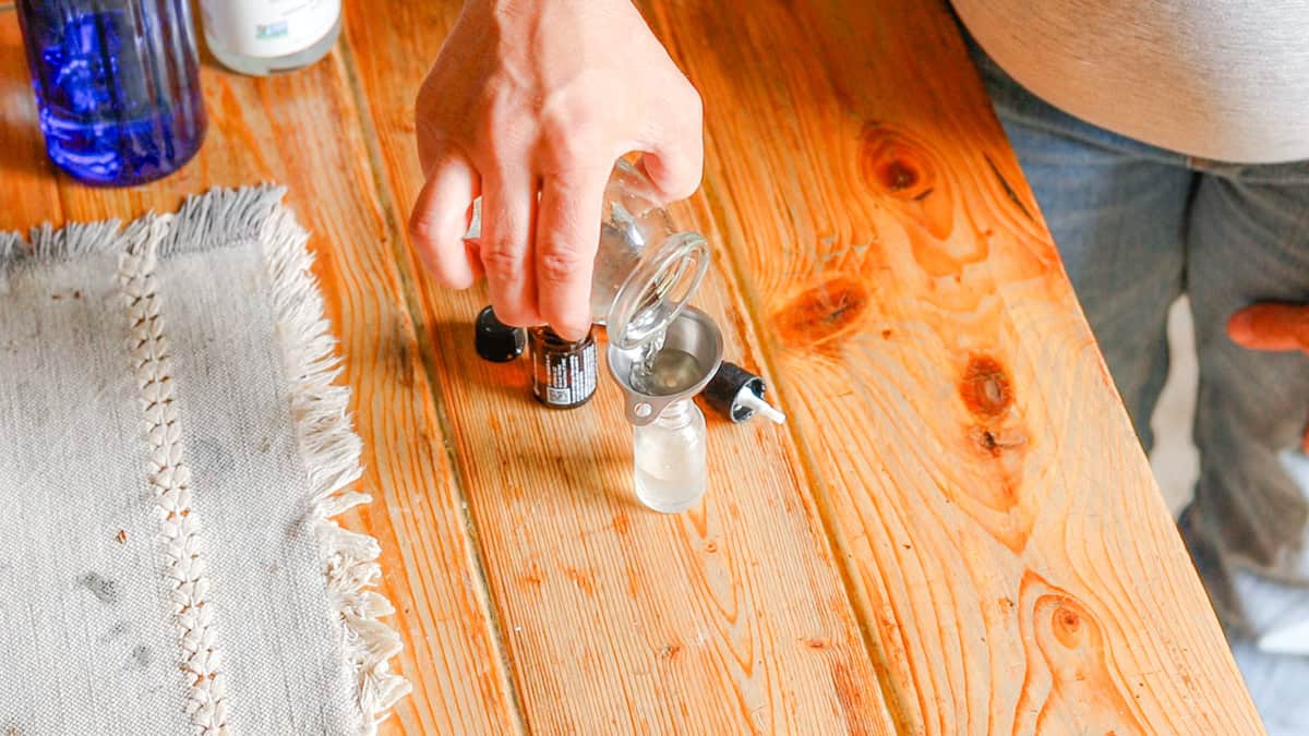 Glycerine is poured into a glass jar as part of a homemade ylang ylang spray perfume recipe. A hand is dropping essential oils into a glass jar for a homemade perfume recipe. The glass jar sits on a wooden table with a linen table runner.