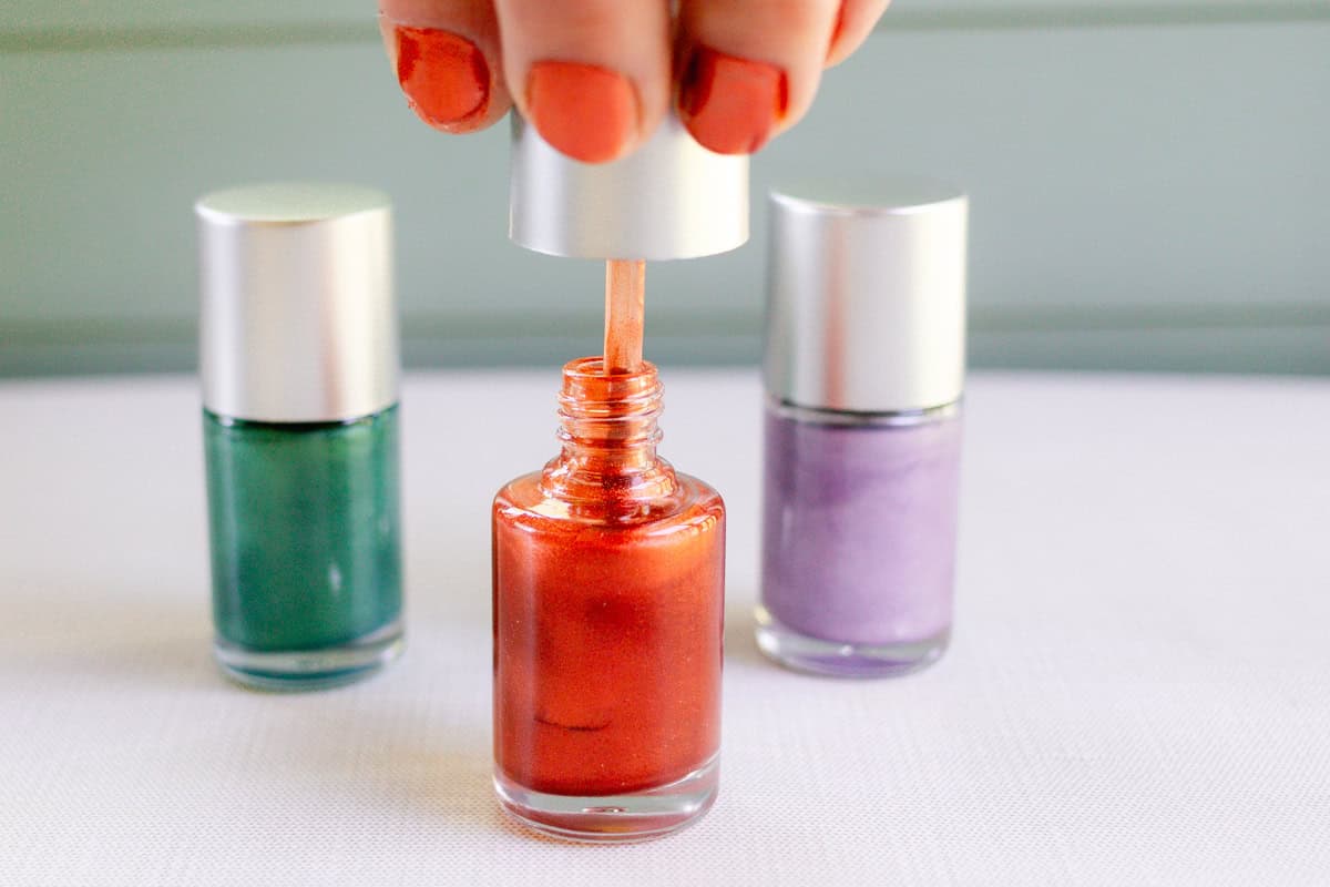 A hand with red nails pulling the brush out of a bottle of homemade red nail polish. A green bottle and a purple bottle of homemade nail polish are in the background.