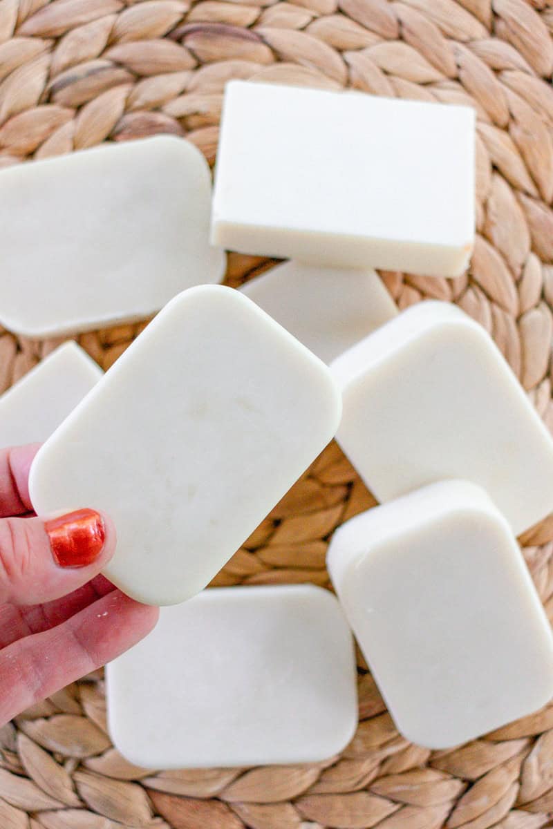 A hand with orange fingernail polish holding up a bar of white cold process facial soap over a rattan placemat. There are 8 rectangular bars of soap pictured. 