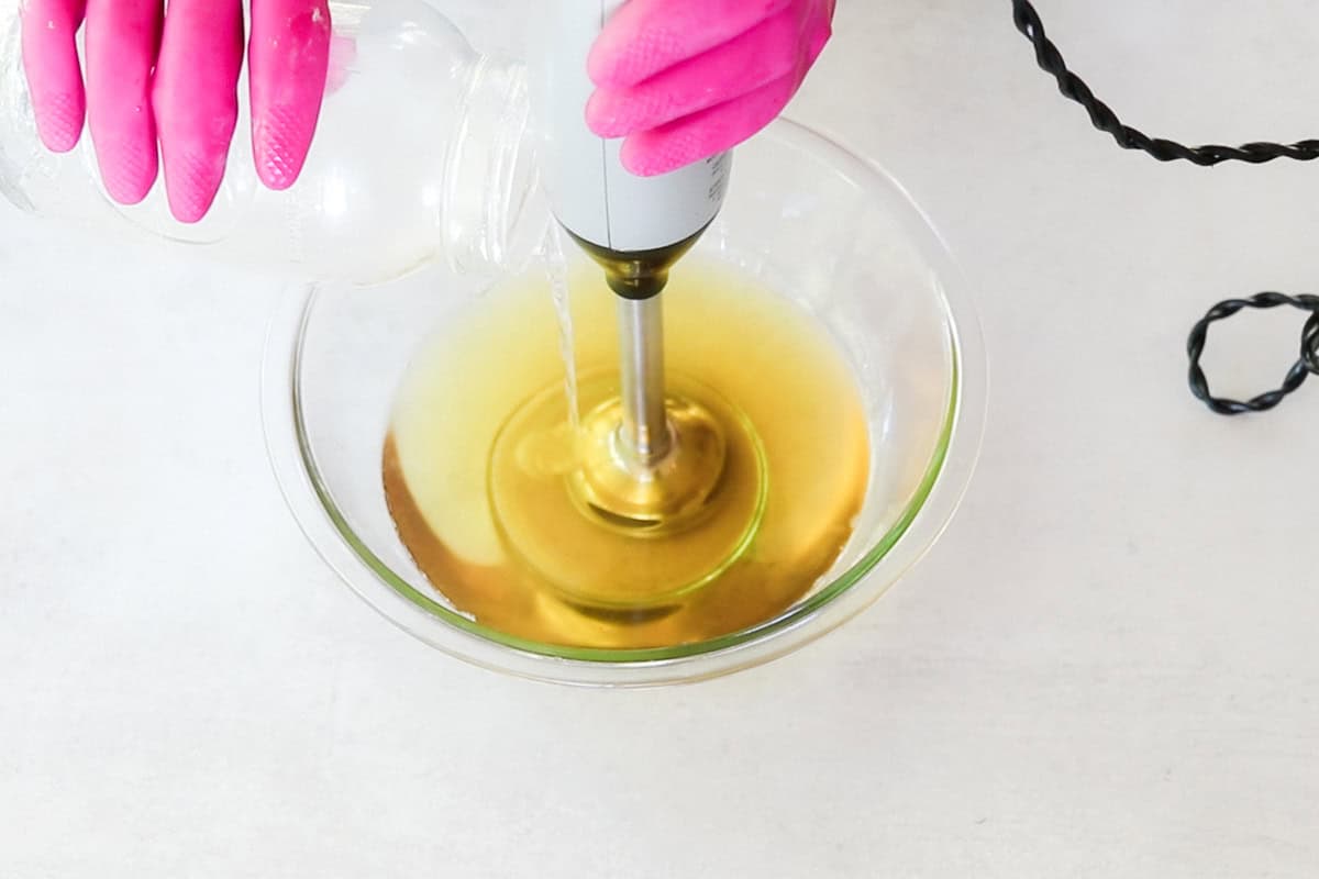 Two hands wearing bright pink rubber gloves use an immersion blender and pour the lye solution into a clear glass bowl filled with melted oils as part of a cold-process facial soap recipe.