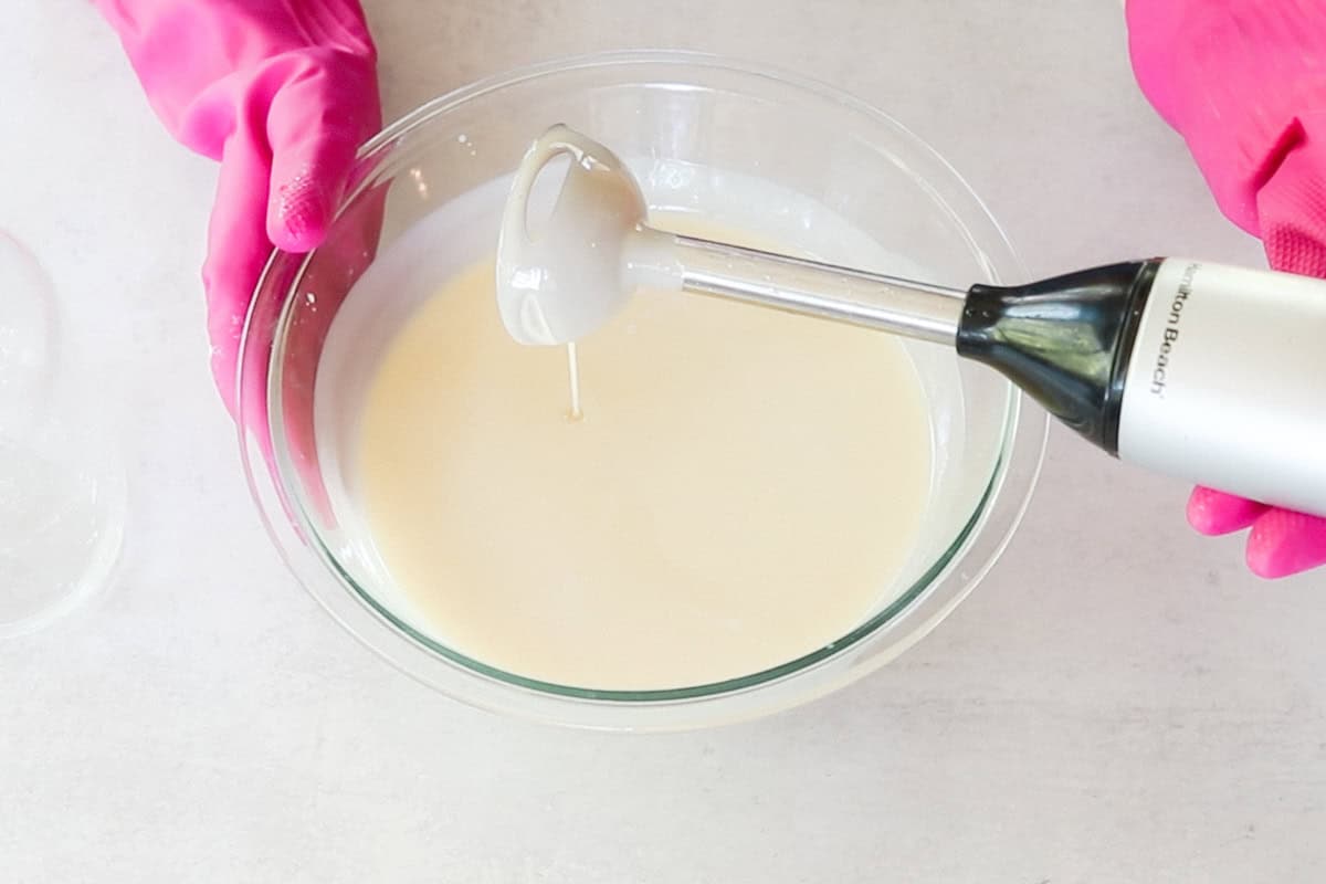 Two hands wearing bright pink rubber gloves use an immersion blender to blend the lye solution with melted oils as part of a cold-process facial soap recipe.