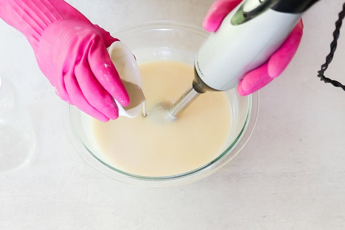 Two hands wearing bright pink rubber gloves use an immersion blender to blend as they add bentonite clay to melted oils and lye as part of a cold-process facial soap recipe.