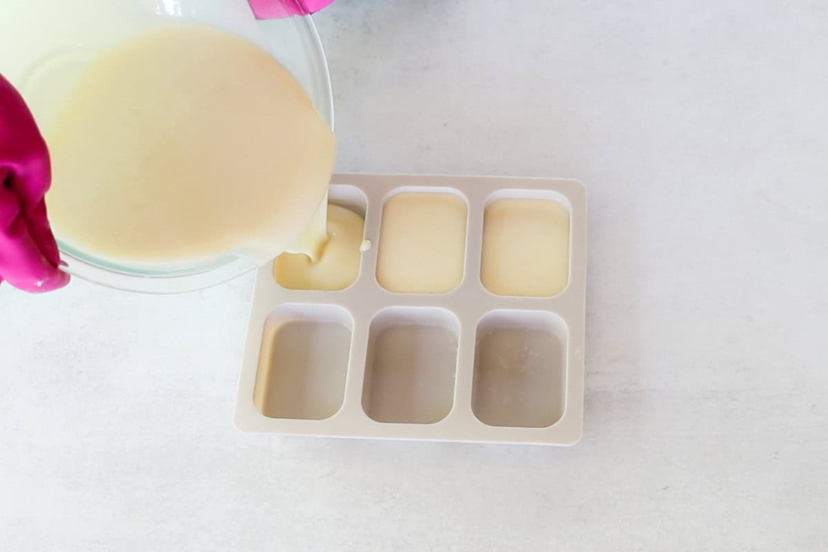 Two hands wearing bright pink rubber gloves pour a mixture into soap molds as part of a cold-process facial soap recipe.