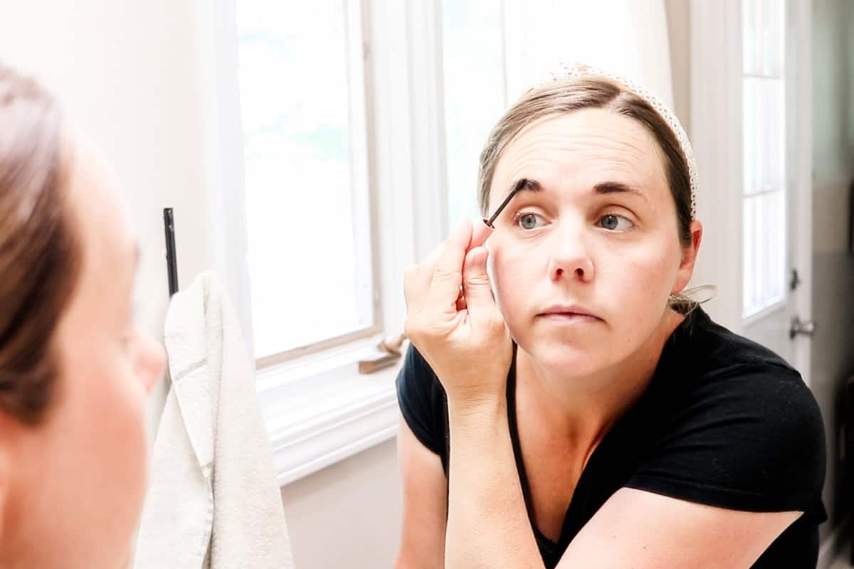 Applying DIY eyebrow tint to my eyebrows in a bathroom mirror with an old mascara brush. 