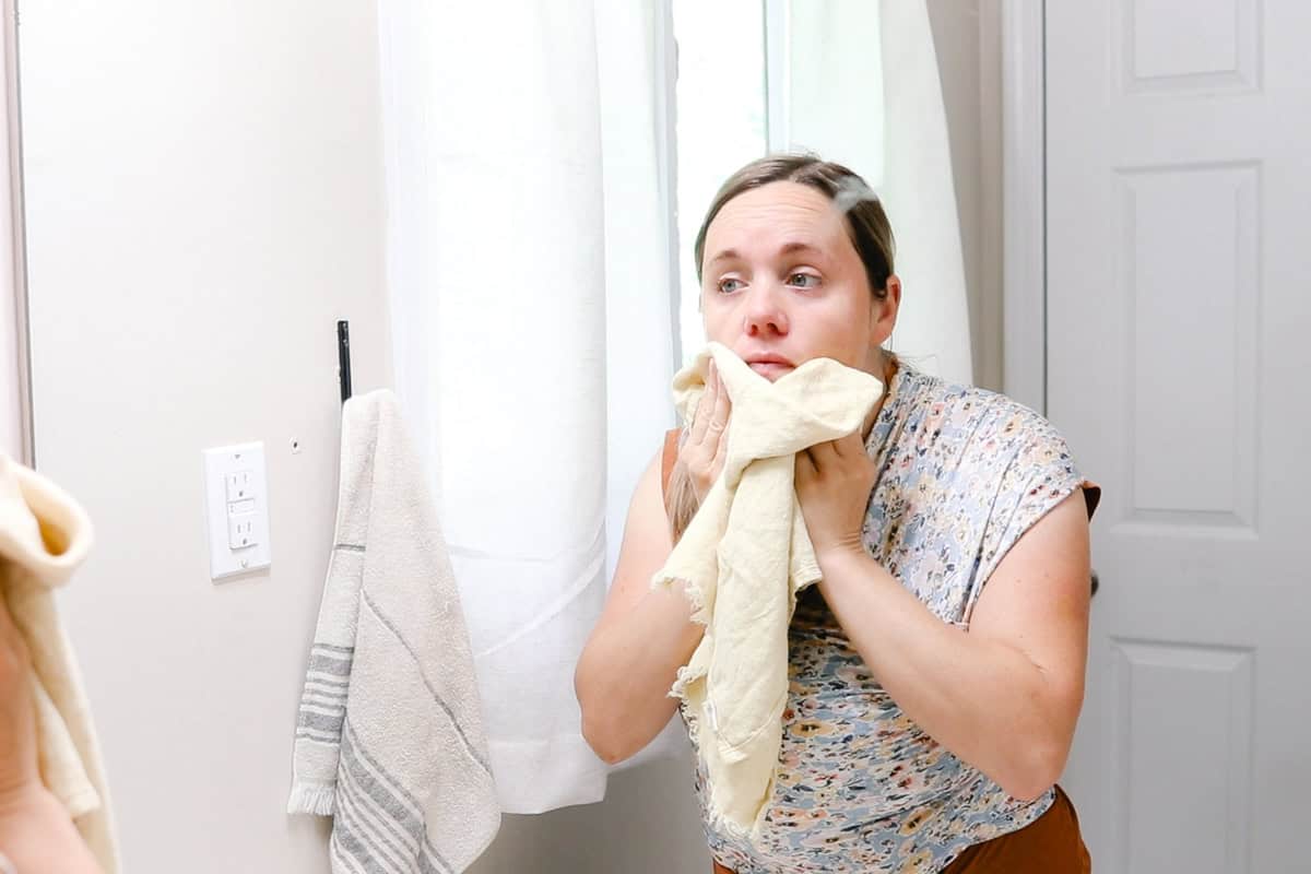 Laura patting her face dry with a towel after cleansing it in a bathroom mirror. 