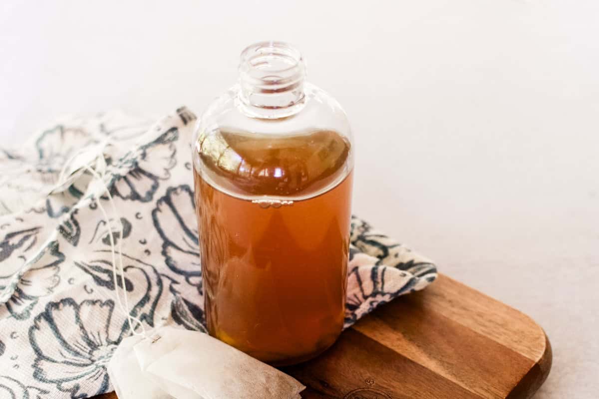 A clear glass bottle of green tea toner sitting next to a tea bag on a wooden cutting board. 
