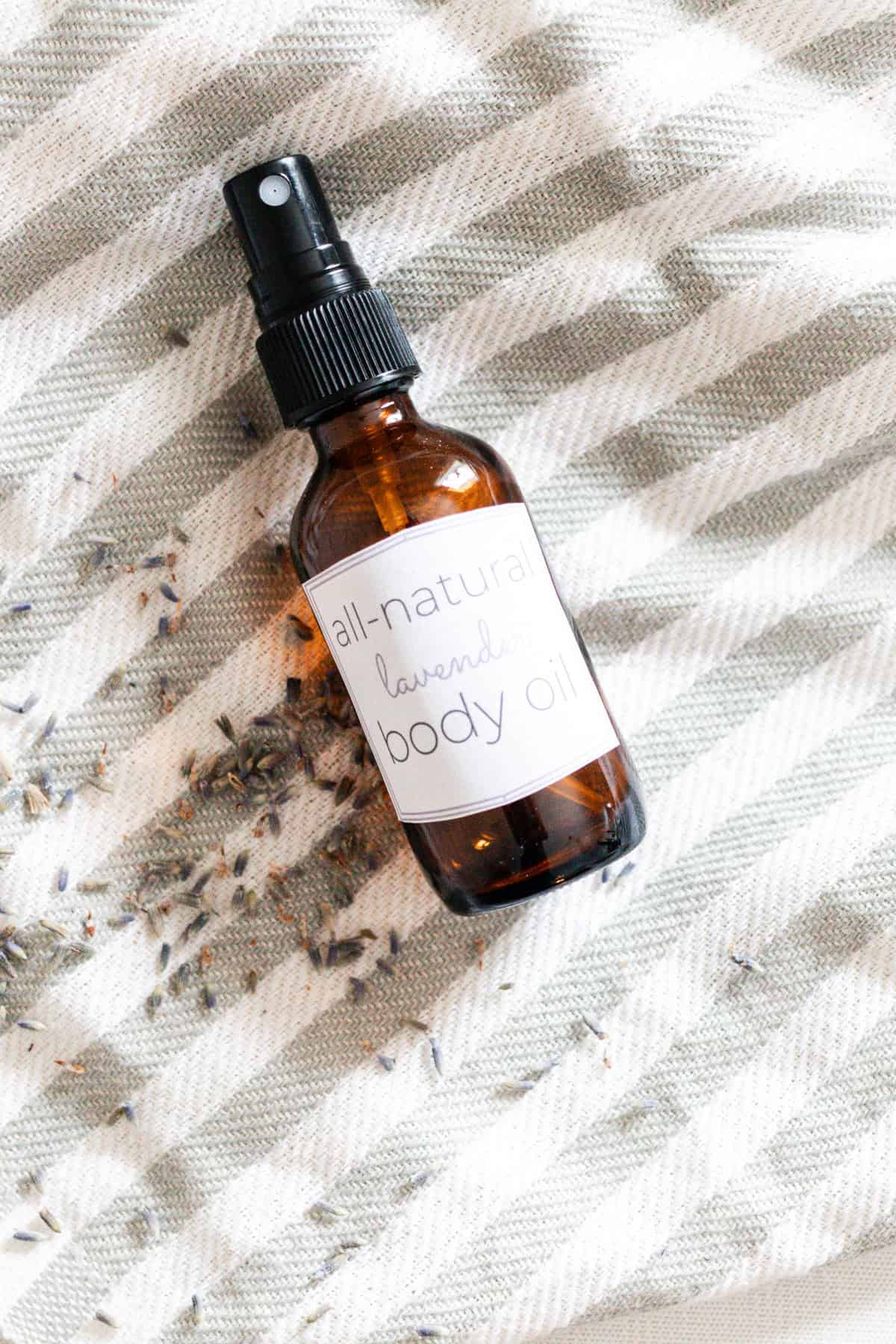 A bottle of homemade body oil laying on a striped towel with dried lavender buds.