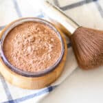 Foundation in a storage container with a makeup brush next to it.