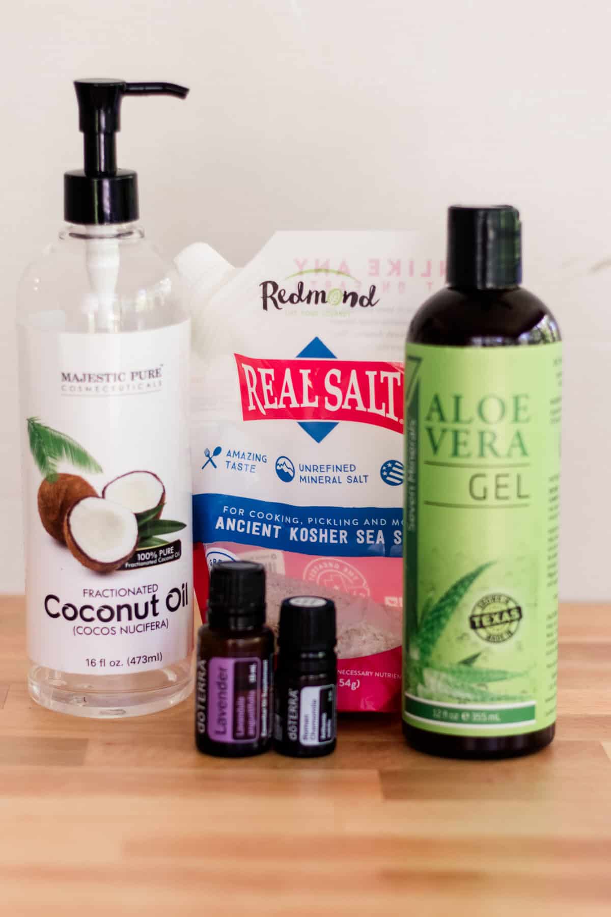 A bottle of coconut oil, a bottle of aloe vera, a bag of sea salt, and two bottles of essential oils assembled on a countertop. 