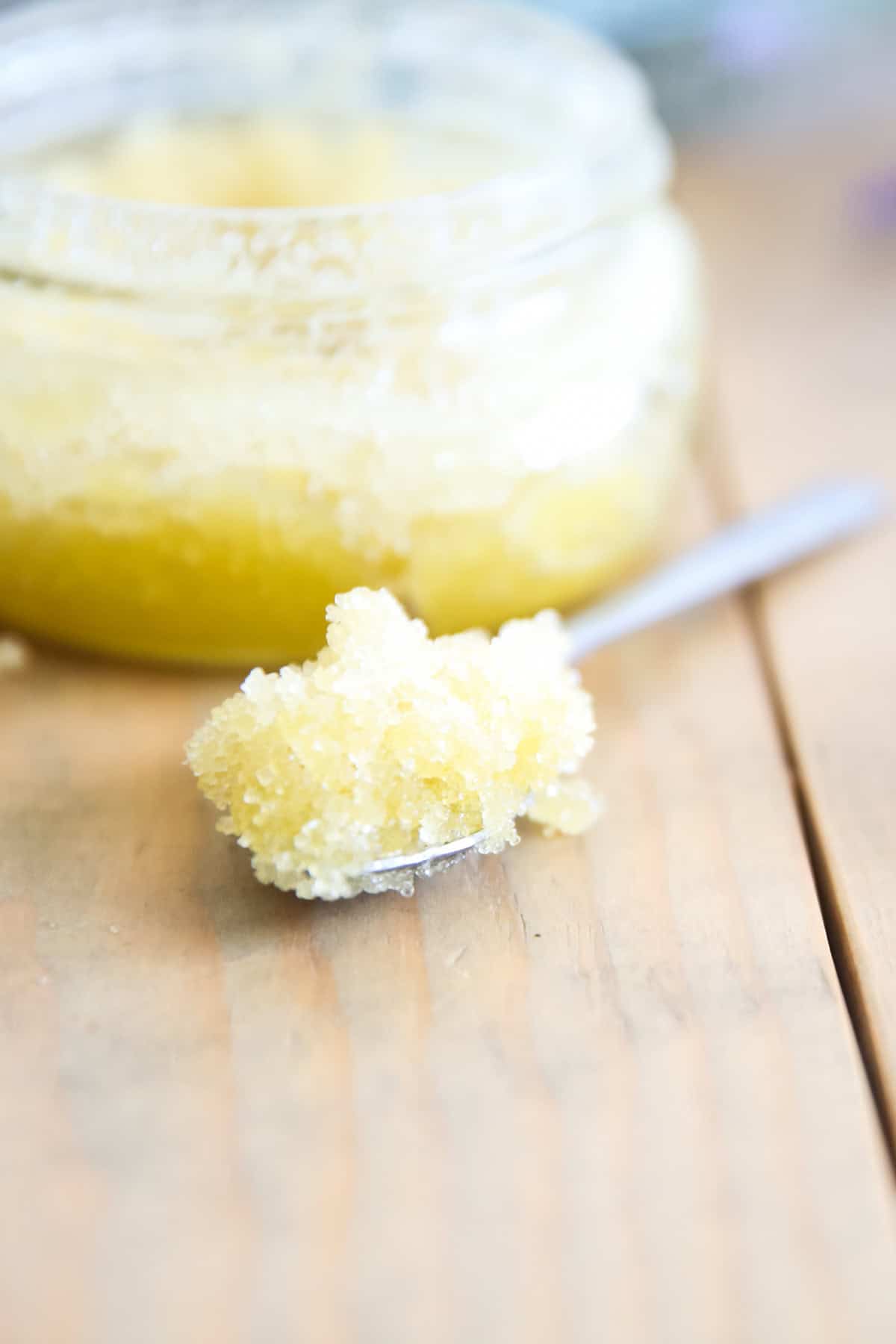 A spoonful of homemade sugar scrub in a spoon with a jar of sugar scrub in the background. 