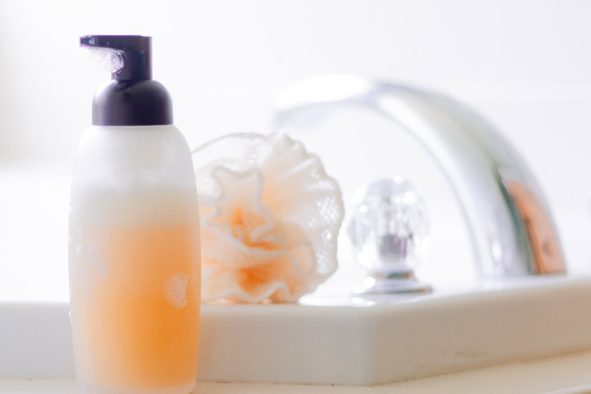 A bottle of natural body wash with a loofah and sink in the background.