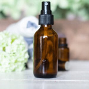 An amber spray bottle of spray deodorant with green flowers, essential oils, and towels in the background.