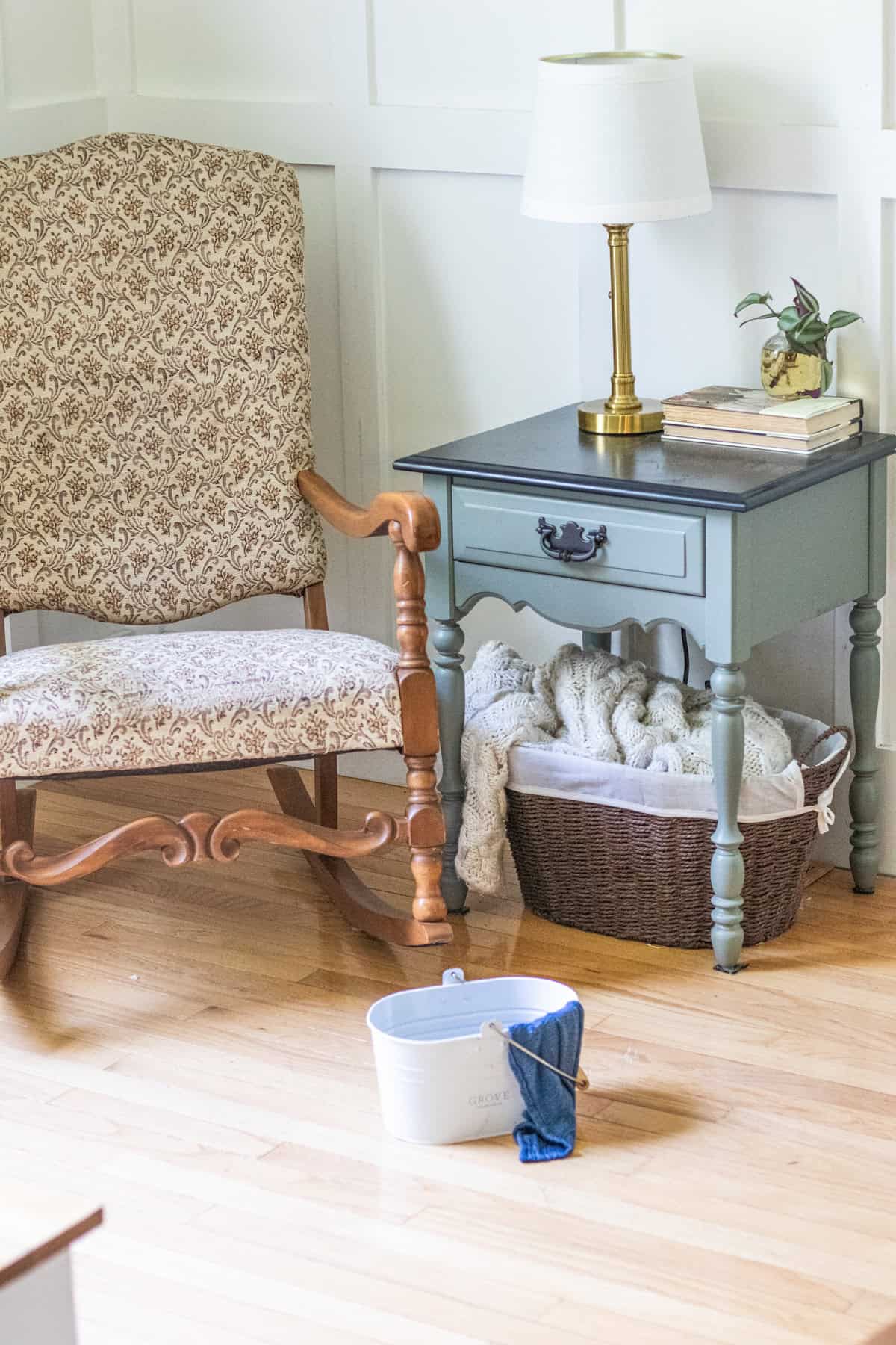 A living room setting with wood floors, a chair, and a side table. A bucket of wood floor cleaner is sitting in the middle of the room.