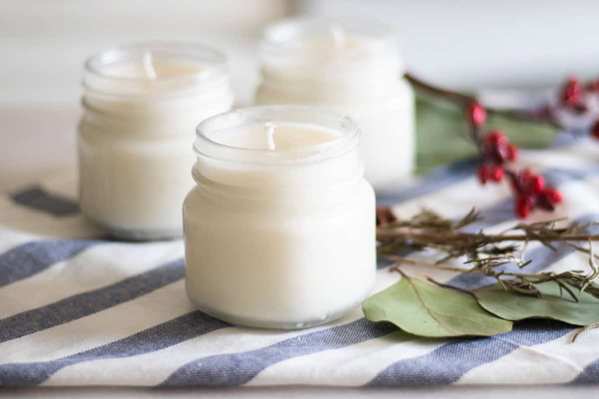 Three homemade Christmas candles with greenery and berries in the background.