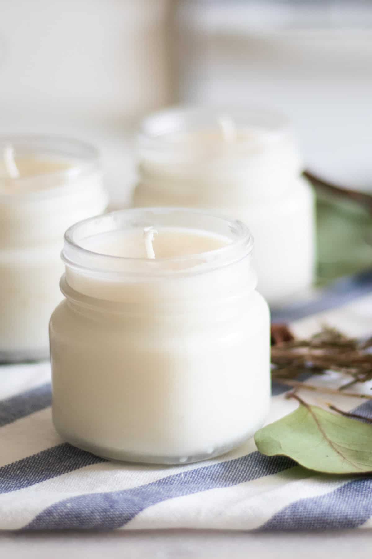 Three homemade Christmas candles with greenery on a white and blue towel.