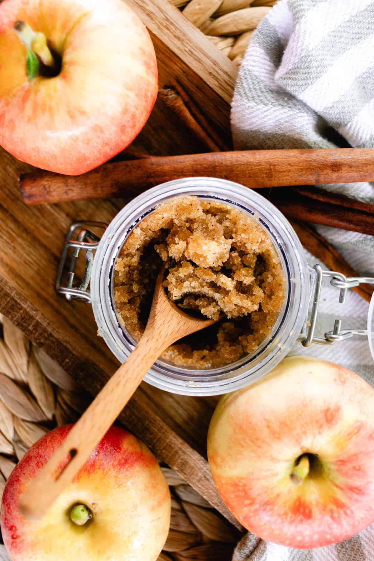 A jar of apple pie body scrub with fresh apples and cinnamon sticks.