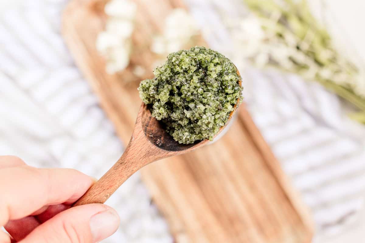 A spoonful of matcha green tea scrub being help up over a wooden cutting board.