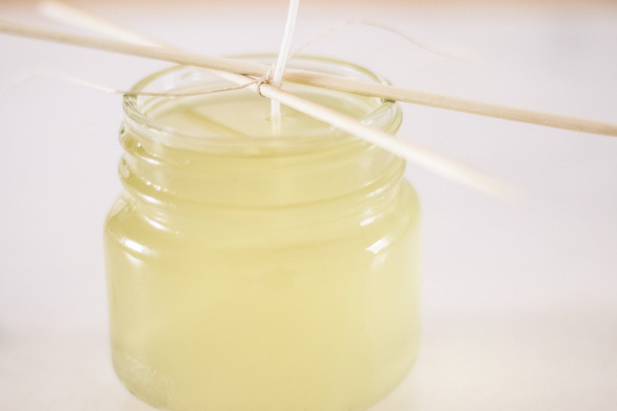 A jar of melted wax with a wick sticking out of it, wrapped around a stick to hold it up.