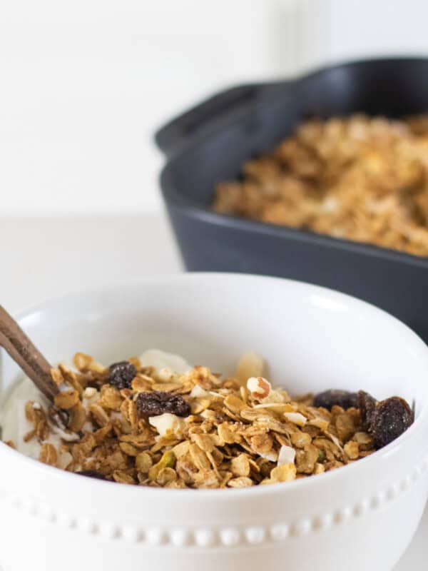 A white bowl filled with honey granola with a baking dish of granola in the background.