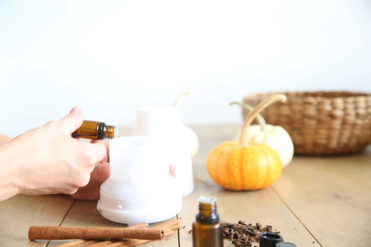 Dropping essential oils into a diffuser that is sitting next to pumpkins, cinnamon sticks, and cloves.