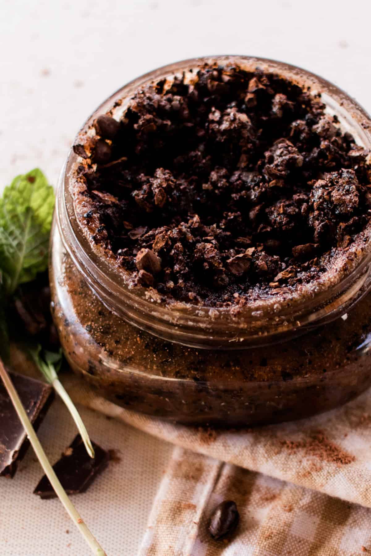 Face scrub spilling out of the top of the storage jar while its sitting on top of a tea towel with fresh peppermint leaves behind it. 