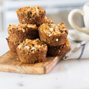 Applesauce muffins stacked on a wooden cutting board to cool with a cup of hot coffee in the background.