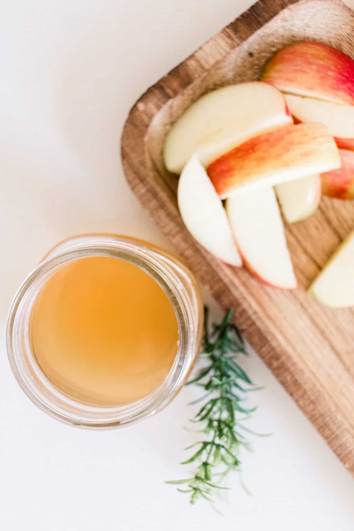 Apple cider vinegar shampoo, red apple slices, and sprigs of rosemary on white table. 