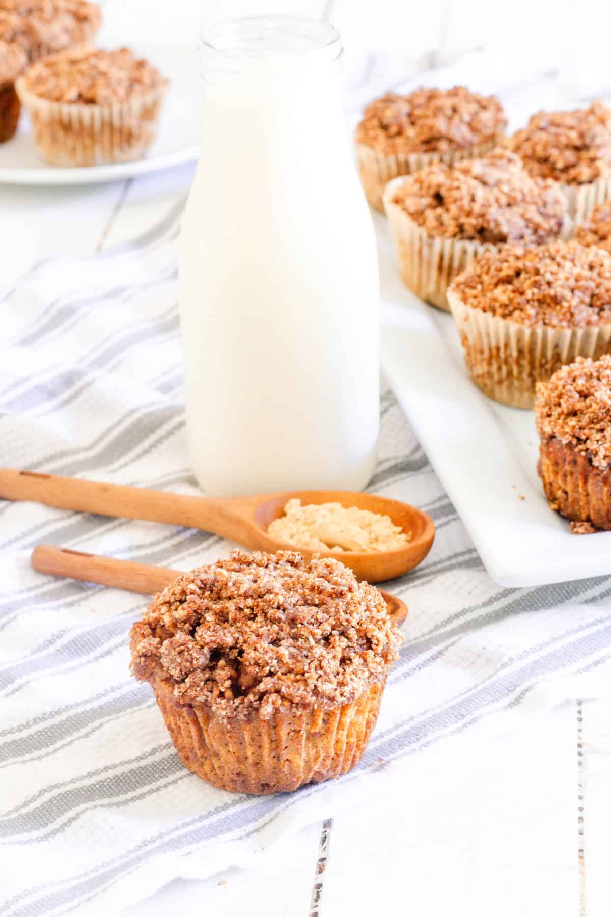 Healthy pumpkin muffins on a light colored towel with a glass milk jar behind it and several more pumpkin muffins on a serving plate. 