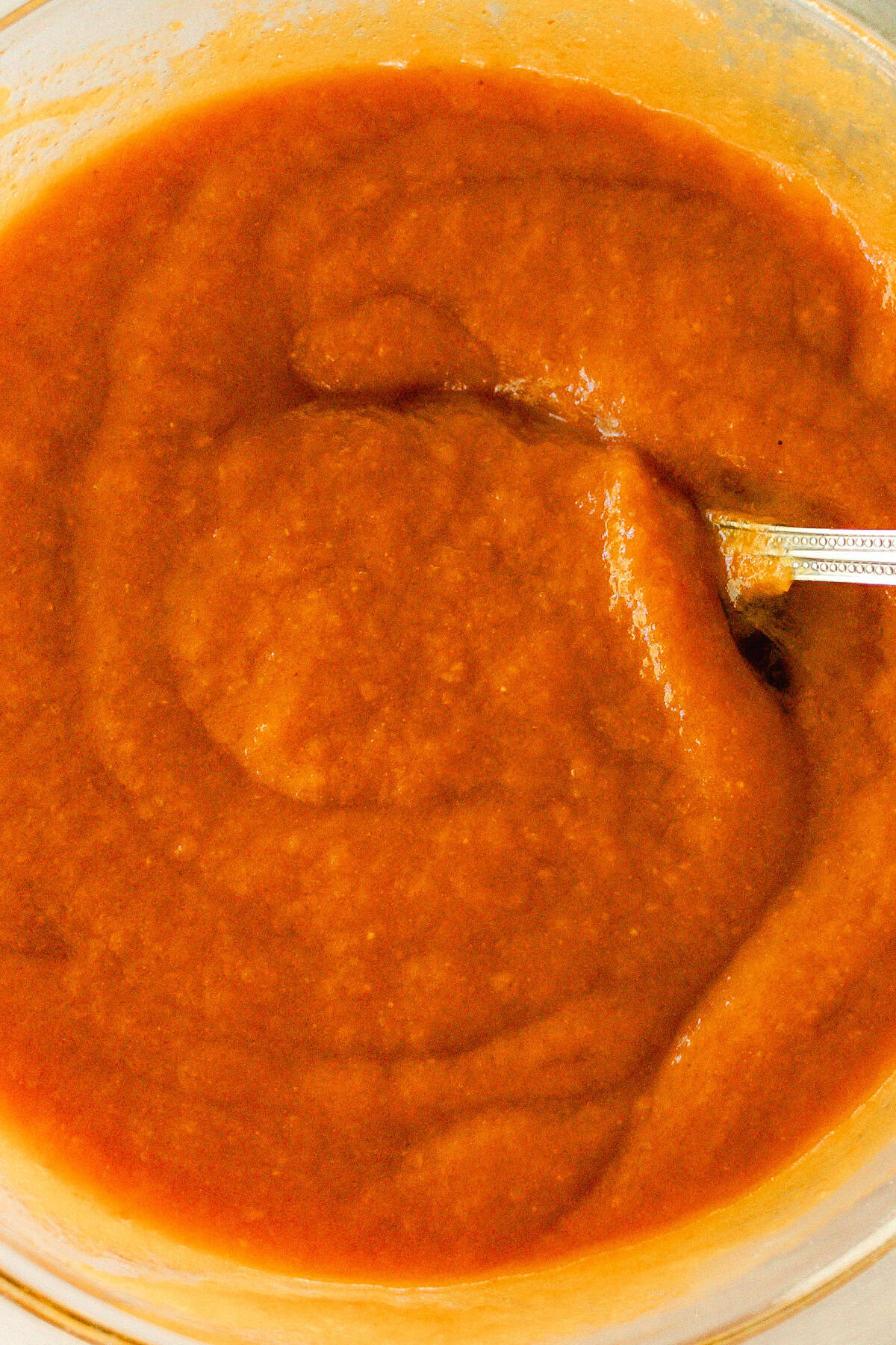 Wet ingredients for the pumpkin muffins getting stirred in a clear mixing bowl with a metal spoon. 