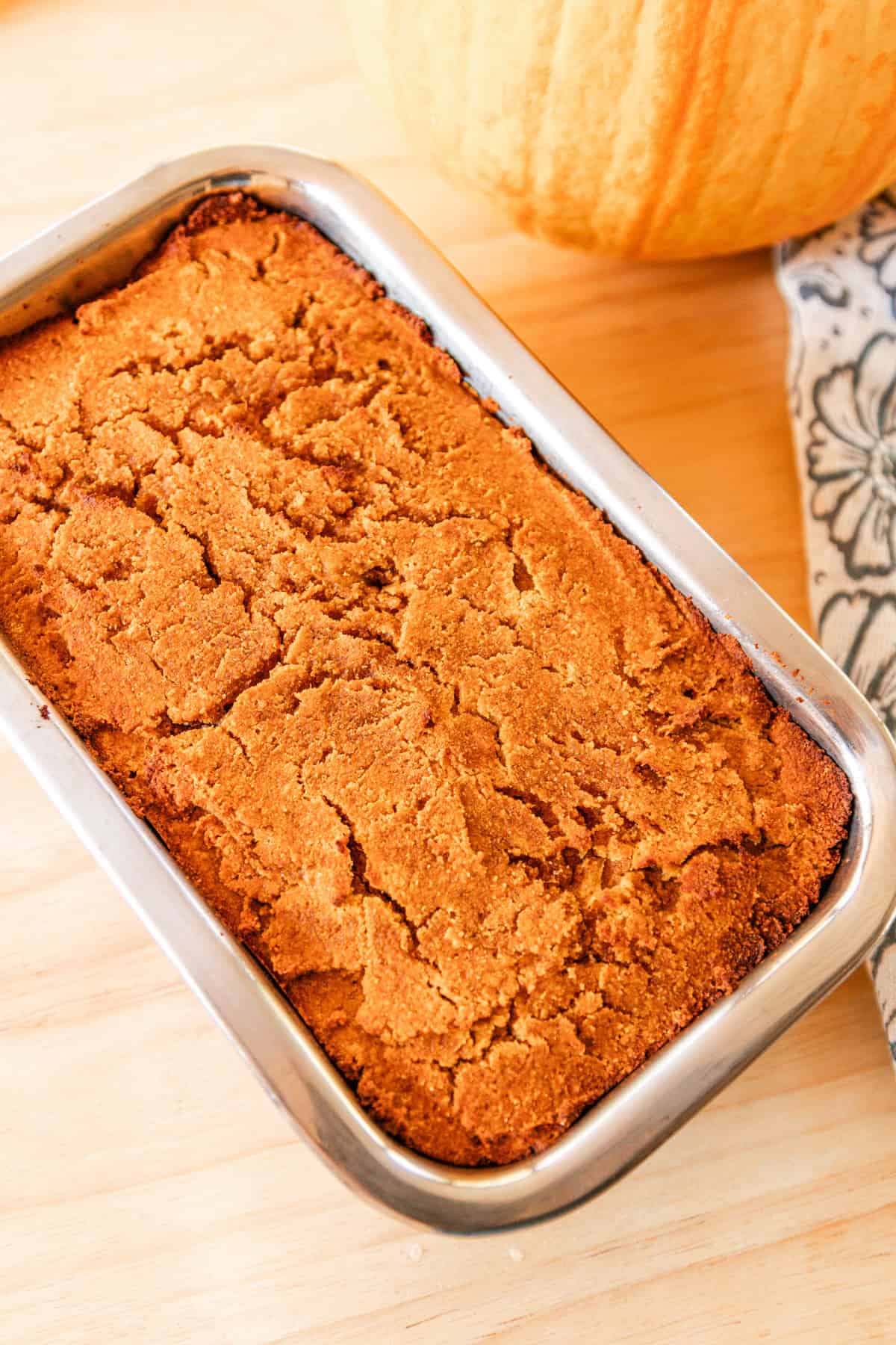 Paleo pumpkin bread cooling in a loaf pan on a wooden table with a decorative flower towel next to it. 
