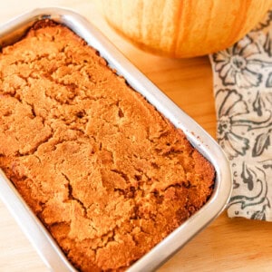 Paleo pumpkin bread cooling in a loaf pan on a wooden tray with a small pie pumpkin next to it.