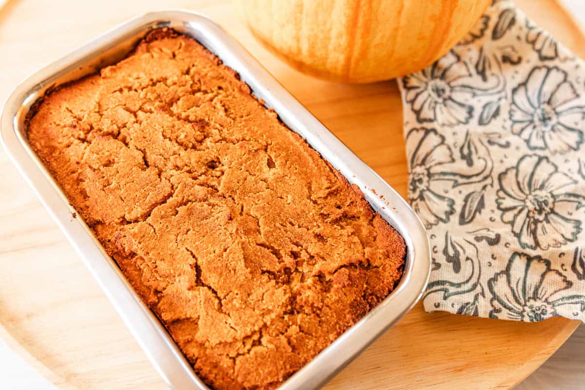 Paleo Pumpkin Bread in loaf pan on a wooden tray with a small, pie pumpkin in the background.