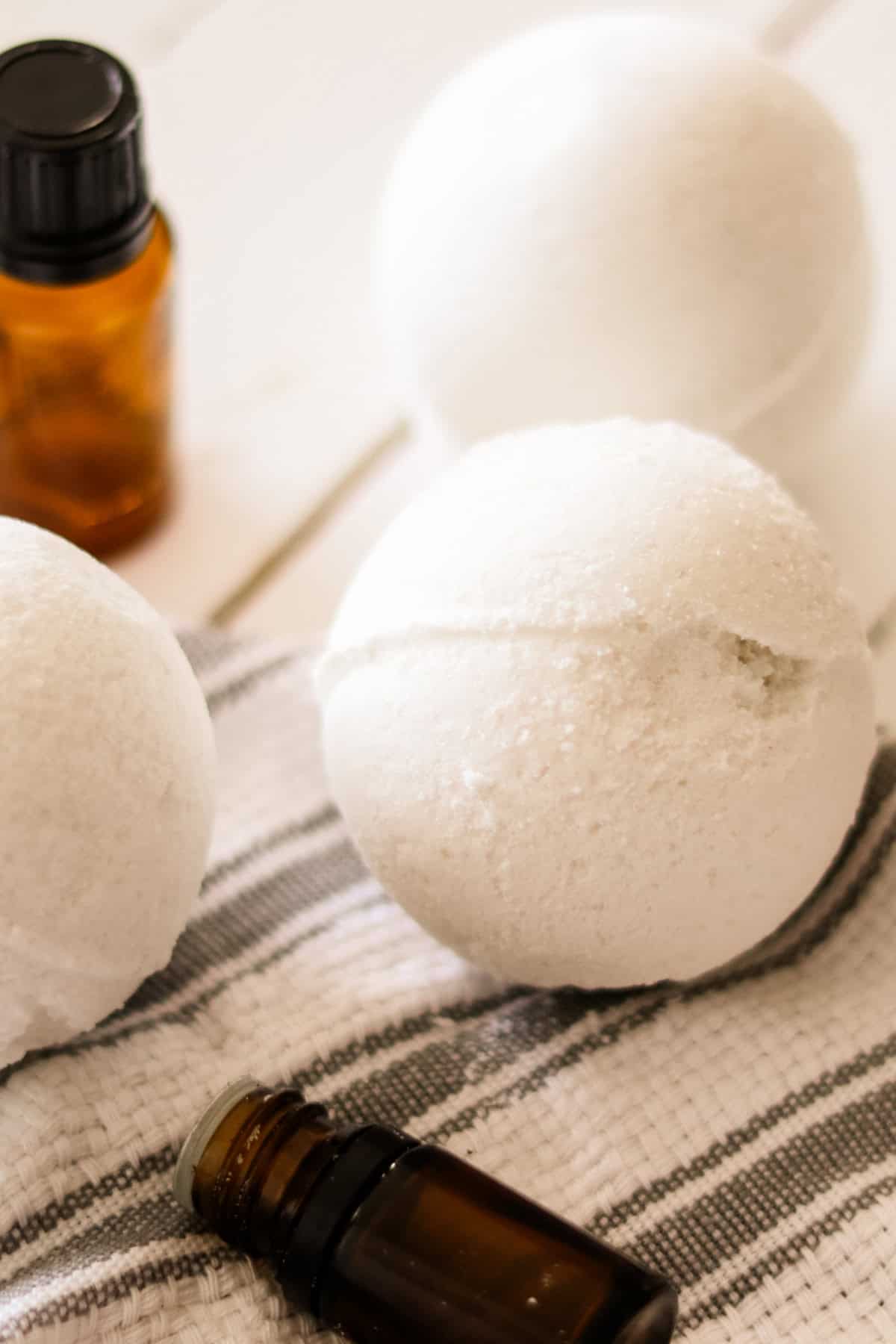 3 homemade bath bombs on a gray and white tea towel with 2 essential oil bottles. 