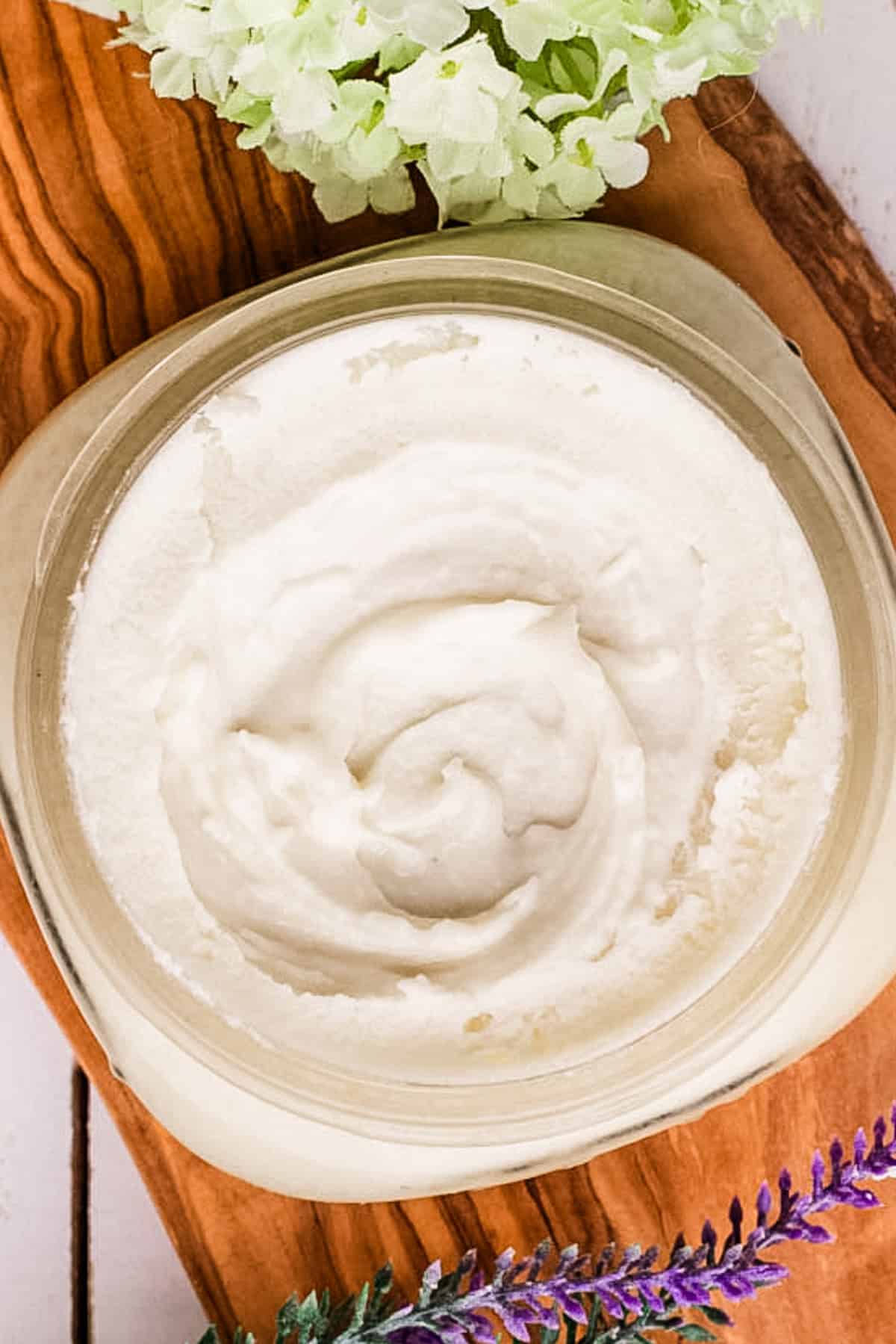 Homemade face moisturizer in a shallow clear container on a wooden tray with lavender sprigs. 