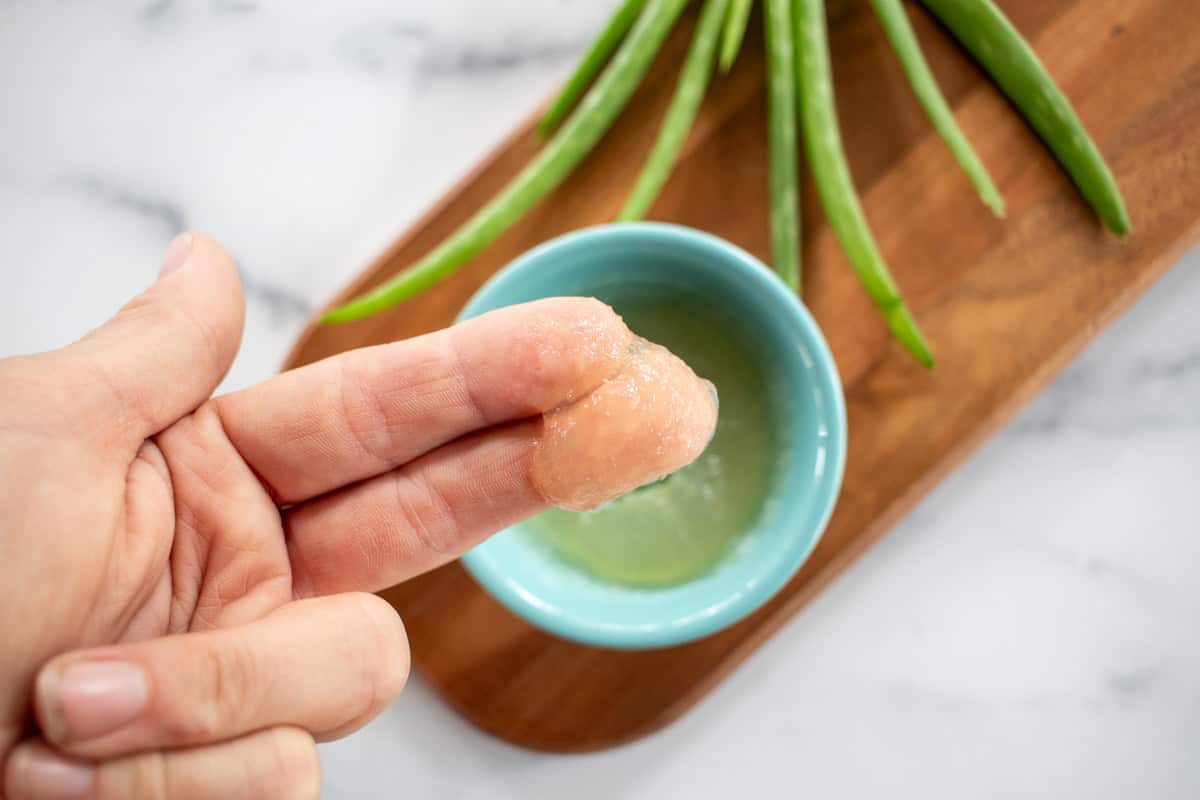 Two fingers with a scoop of aloe vera on them over a bowl of aloe vera.