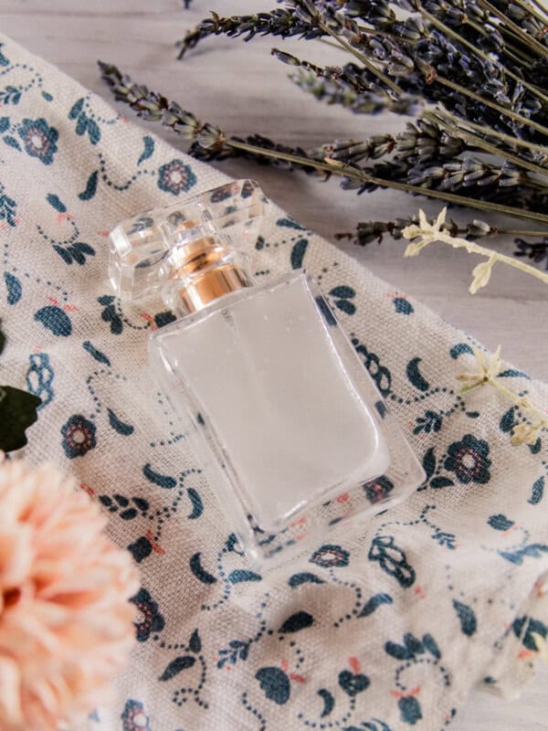 A glass bottle of homemade perfume on a floral towel with flowers around it.