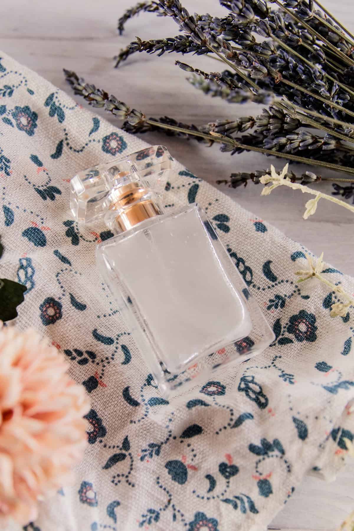 A glass bottle of homemade perfume on a floral towel with flowers around it.