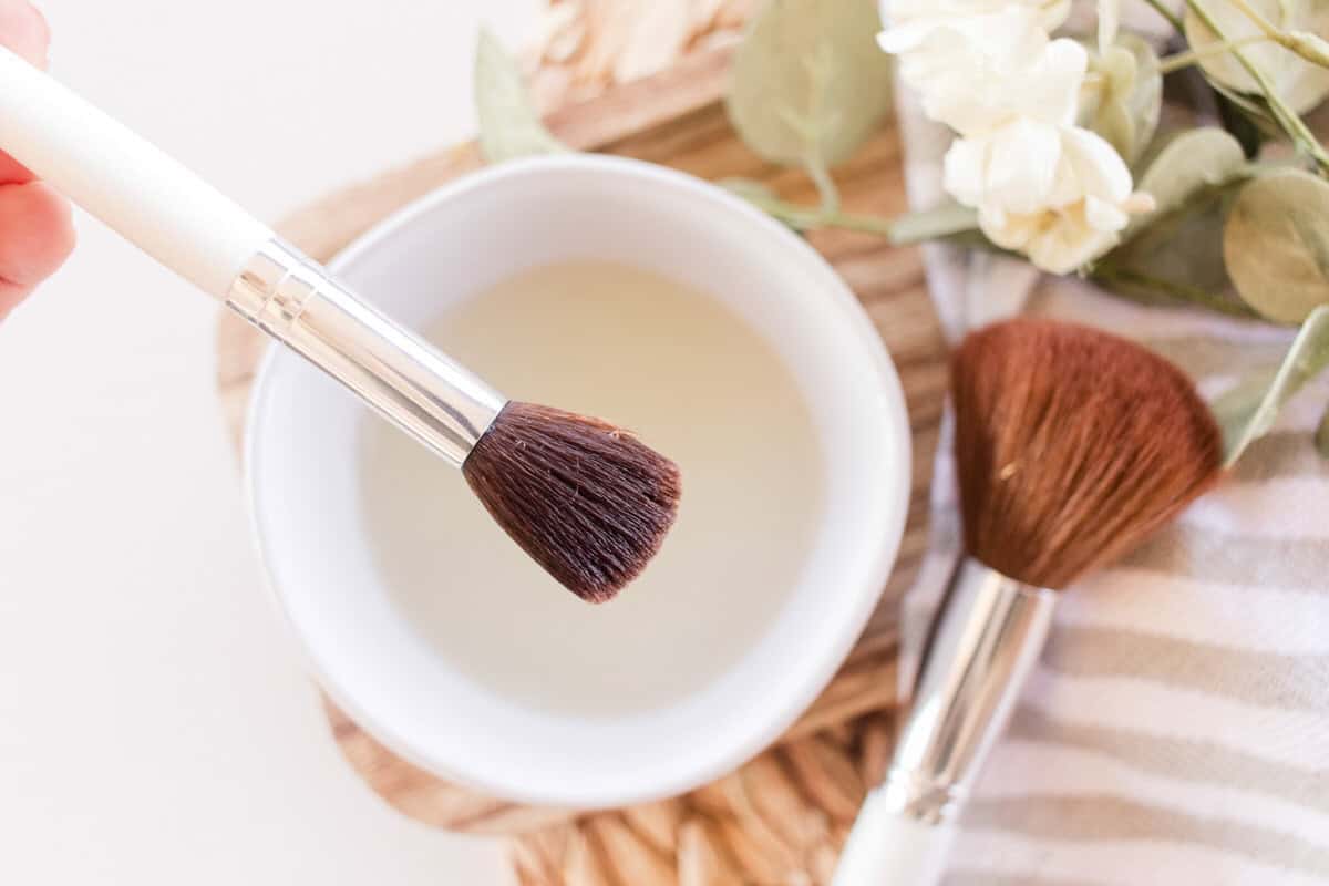 Holding a makeup brush over a bowl of homemade makeup cleaner.