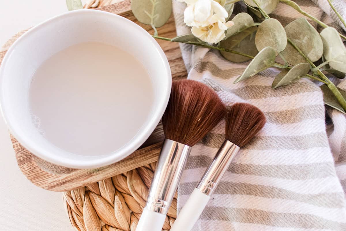 A bowl of makeup brush cleaner with makeup brushes on a striped towel.