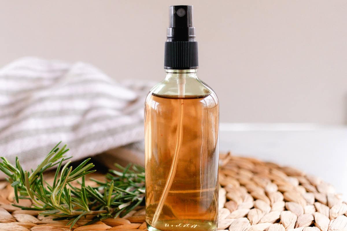 Glass spray bottle of rosemary water on a white table with fresh rosemary springs and a grey and white striped tea towel next to it.