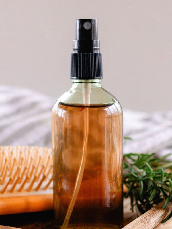 Homemade rosemary water in a clear spray bottle on a wooden top with a comb and fresh sprigs of rosemary next to it.