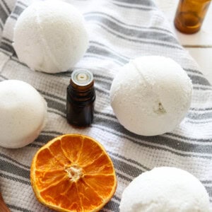 Several homemade bath bombs on a grey and white striped tea towel with dried oranges, essential oil bottles around it.