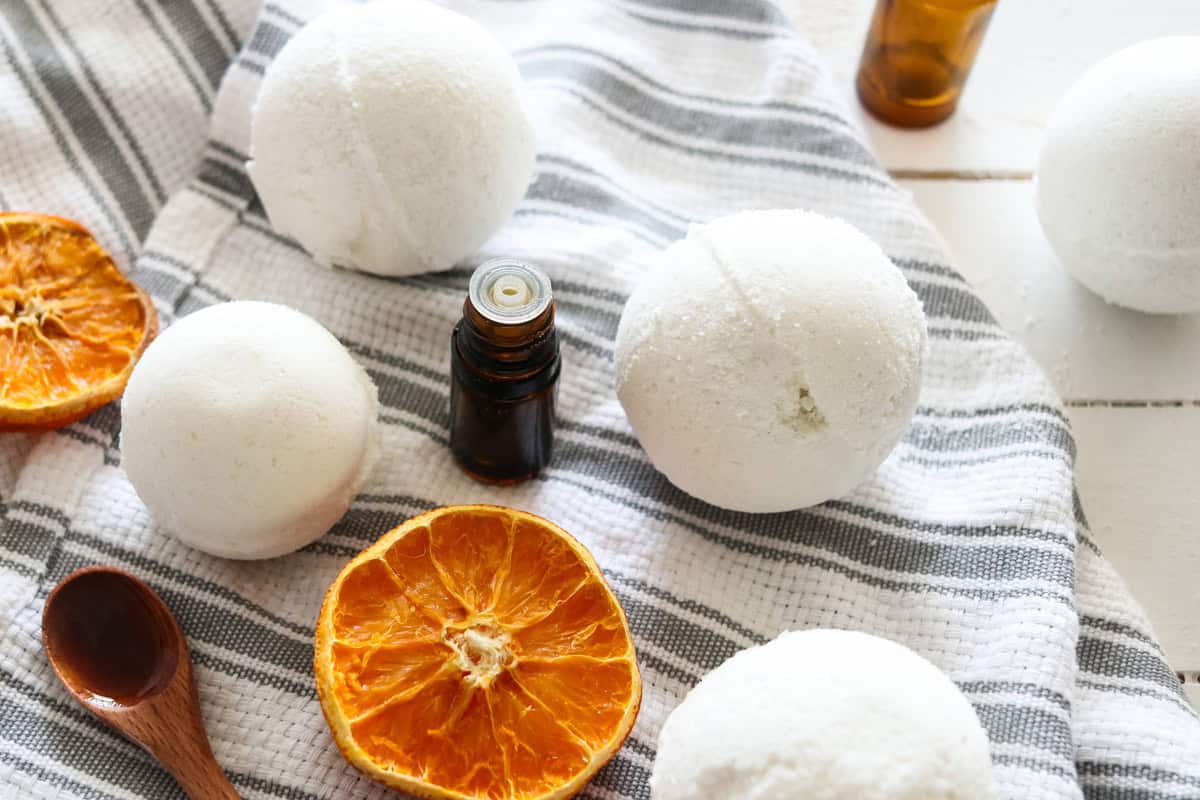 Several homemade bath bombs on a grey and white striped tea towel with dried oranges, essential oil bottles around it.