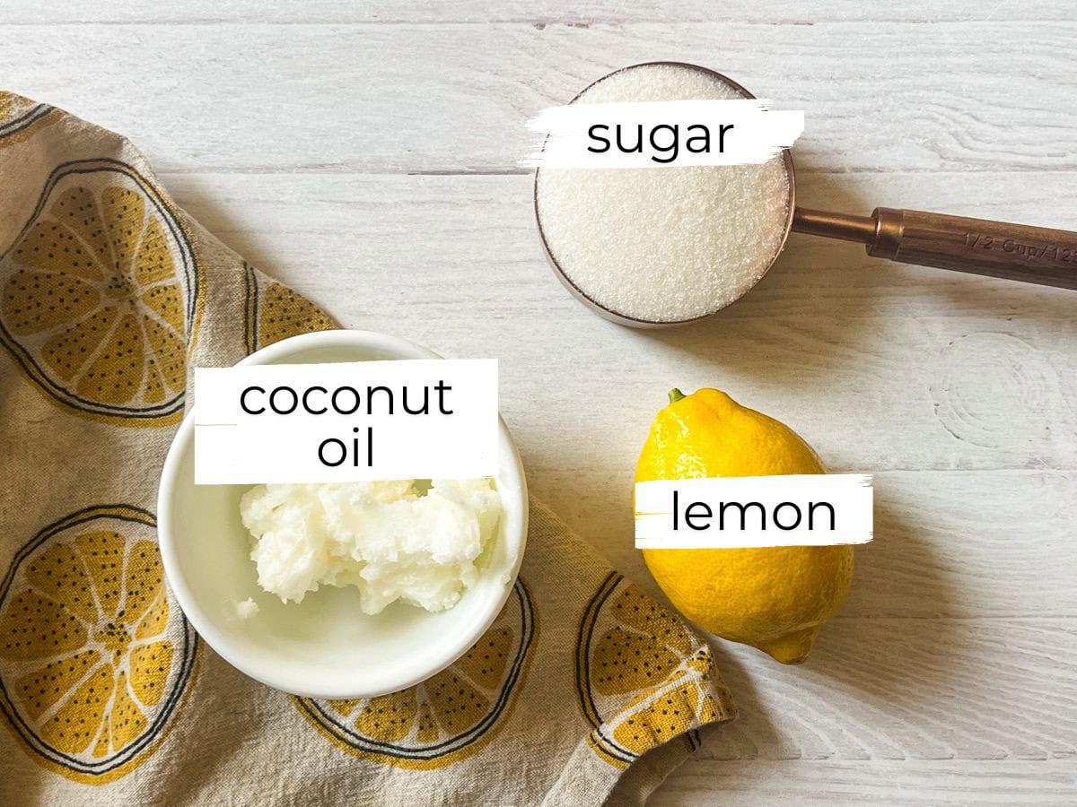 Homemade hand scrub ingredients assembled and labeled on a white table with a lemon tea towel.