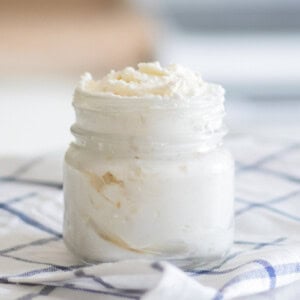 A jar of homemade shea butter lotion on a white and blue checkered tablecloth.