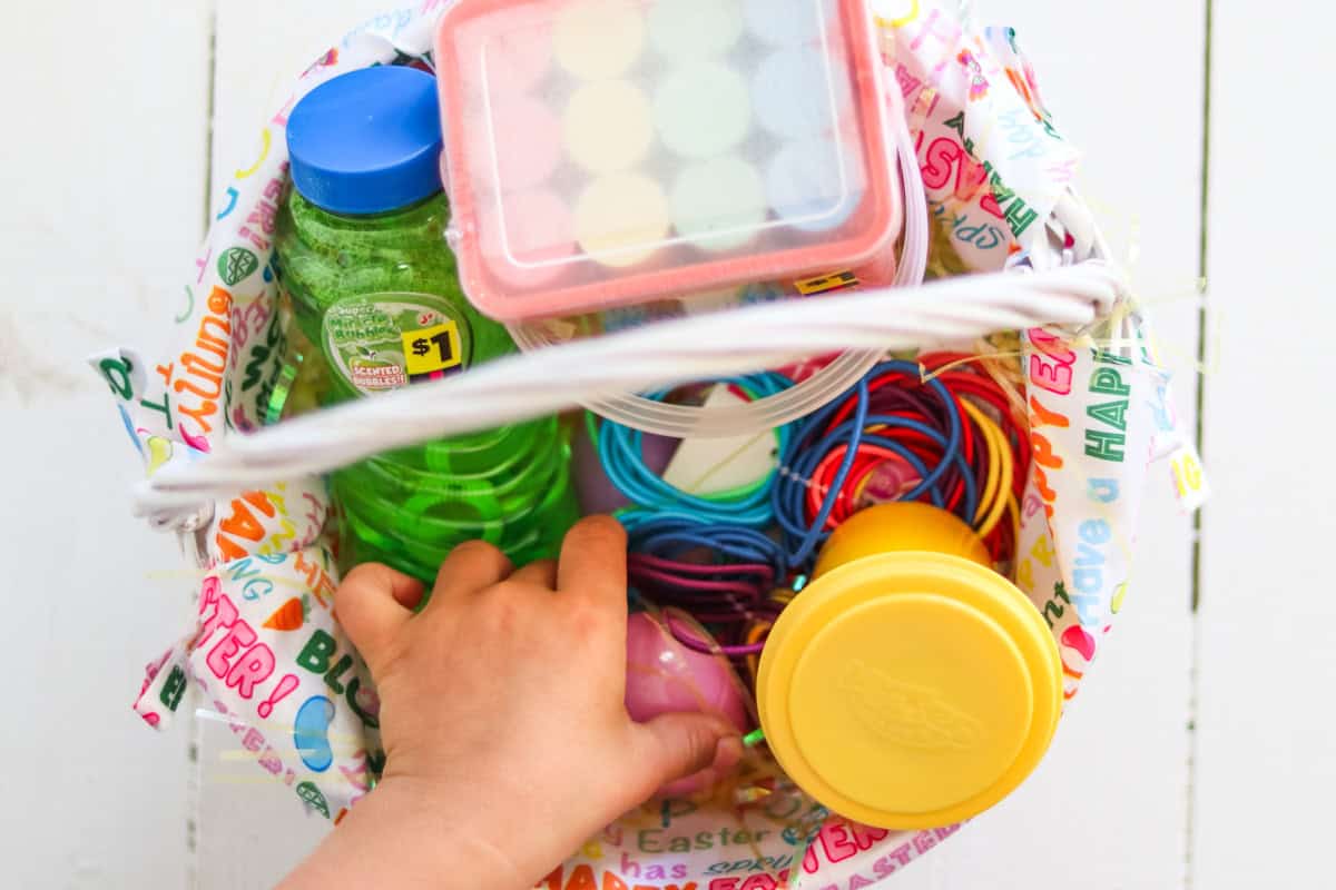 A child's hand reaching into an Easter basket filled with bubbles, chalk, and other toys.